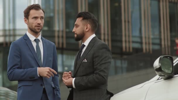 Średnie ujęcie dwóch różnych partnerów biznesowych ubranych w formalne garnitury, stojących samochodem na pierwszym planie nowoczesnego biurowca, uśmiechniętych, mówiących, ściskających dłonie - Materiał filmowy, wideo