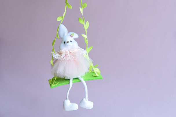 ピンクの背景に卵とイースターの装飾でいっぱいの巣の前で緑の花のスイングで美しい明るいウサギ. - 写真・画像