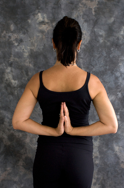 Uma mulher morena caucasiana está de pé em roupas de ioga preto com as mãos atrás das costas em oração reversa
 - Foto, Imagem