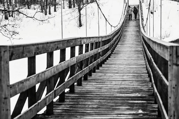 στην ασπρόμαυρη φωτογραφία απεικονίζονται δύο έφηβοι να περπατούν μαζί δίπλα σε μια παλιά πεζοδρομημένη ξύλινη γέφυρα και να πέφτει ελαφρύ χιόνι - Φωτογραφία, εικόνα