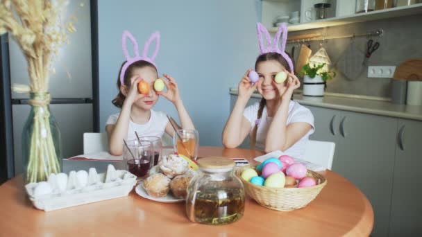 Hyvää pääsiäistä. Kaksi siskoa maalaa pääsiäismunia. Onnelliset perheen lapset valmistautuvat pääsiäiseen. Söpö pieni lapsi tyttö yllään pupu korvat poseeraa värillisiä munia kameran edessä - Materiaali, video