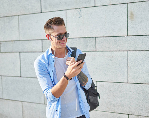 Ein junger Mann, europäisches Aussehen, mit Sonnenbrille und schwarzem Rucksack. Steht an der Wand, schaut ins Telefon und lächelt. - Foto, Bild