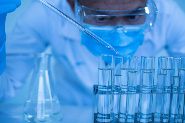 Koruyucu gözlük takan ve eldiven takan bir erkek bilim adamı aşıyı test etmek için kimyasallar damlatıyor. Biyokimyagerler yeni ilaçlar keşfetmek için deneyler yapıyorlar. Araştırmacılar bilim laboratuvarlarında çalışırlar.. - Fotoğraf, Görsel