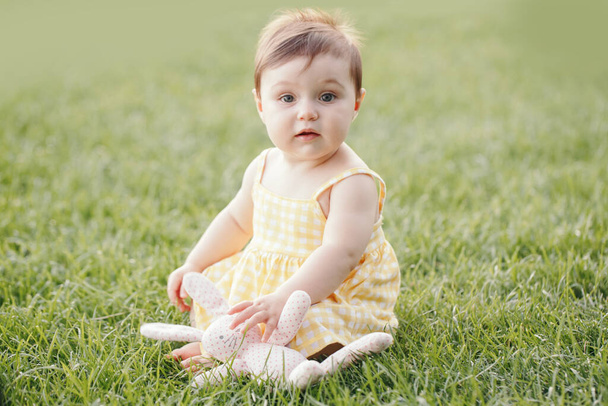Funny baby kid with toy. Cute adorable baby girl in yellow dress sitting on grass in park outdoor. Funny child toddler playing and having fun on summer day. Authentic lifestyle happy childhood.  - Photo, Image