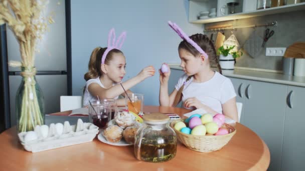 Καλό Πάσχα. Δύο αδερφές βάφουν πασχαλινά αυγά. Ευτυχισμένα παιδιά της οικογένειας ετοιμάζονται για το Πάσχα. Χαριτωμένο κοριτσάκι που φοράει αυτιά κουνελιού. Τα αυγά μαλώνουν στην κάμερα. - Πλάνα, βίντεο