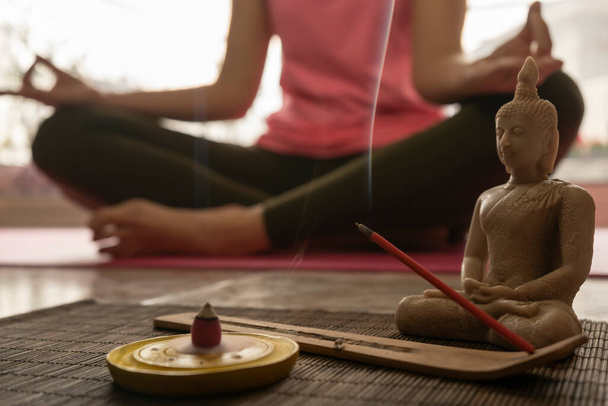 Mladá žena v lotosové pozici meditující doma. Nerozpoznatelná žena praktikující jógu v pozadí se zaměřením na buddhu a kadidlo hořící kouřem. - Fotografie, Obrázek