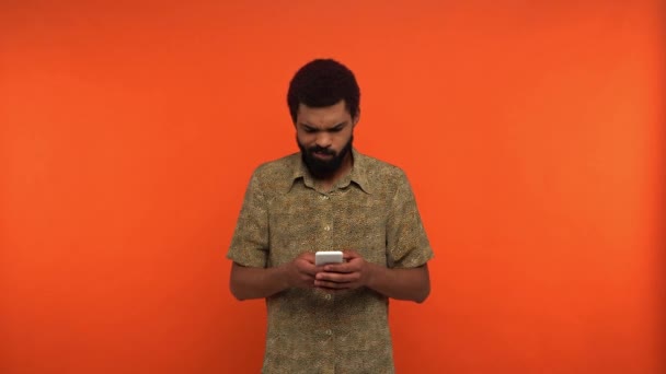 verärgerte afrikanisch-amerikanische Mann Messaging auf Smartphone isoliert auf orange - Filmmaterial, Video