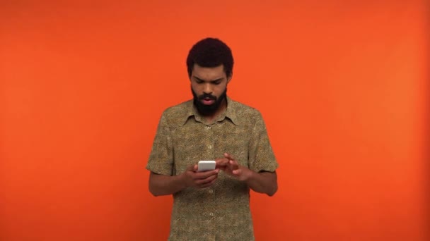 ενθουσιασμένος αφρικάνος Αμερικανός άνθρωπος μηνυμάτων σε smartphone απομονωμένο σε πορτοκαλί - Πλάνα, βίντεο