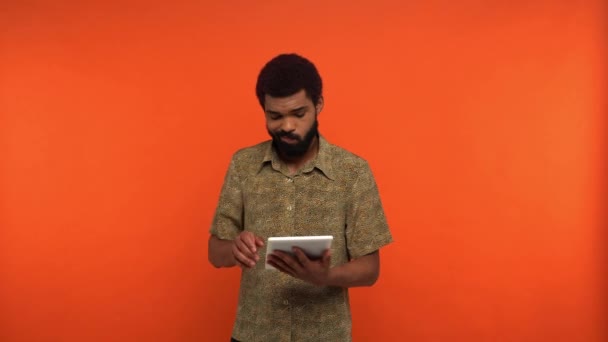 hombre afroamericano insatisfecho usando tableta digital aislada en naranja - Metraje, vídeo