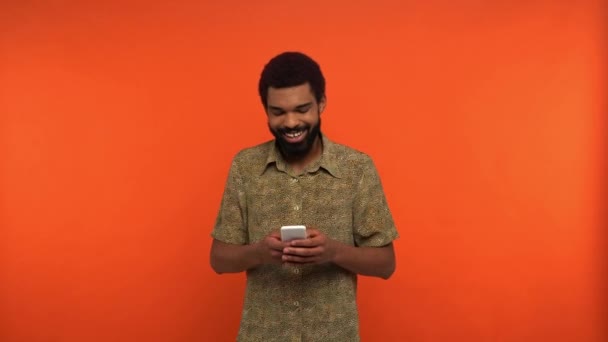 smiling african american man texting on smartphone isolated on orange - Felvétel, videó