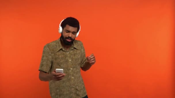 Afrykański Amerykanin w słuchawkach bezprzewodowych przy użyciu smartfona odizolowanego na pomarańczowo - Materiał filmowy, wideo