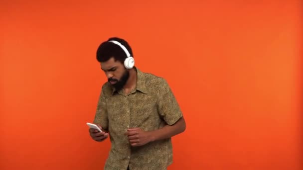 Afrykański Amerykanin w słuchawkach ze smartfonem odizolowanym na pomarańczowo - Materiał filmowy, wideo