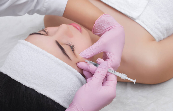 El médico cosmetólogo realiza el procedimiento de inyección de toxina botulínica para apretar y alisar las arrugas en la piel de la cara de una hermosa mujer joven en un salón de belleza.. - Foto, imagen