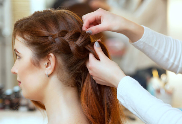 Beautiful, red-haired girl with long hair, hairdresser weaves a French braid, in a beauty salon. Professional hair care and creating hairstyles. - Photo, Image