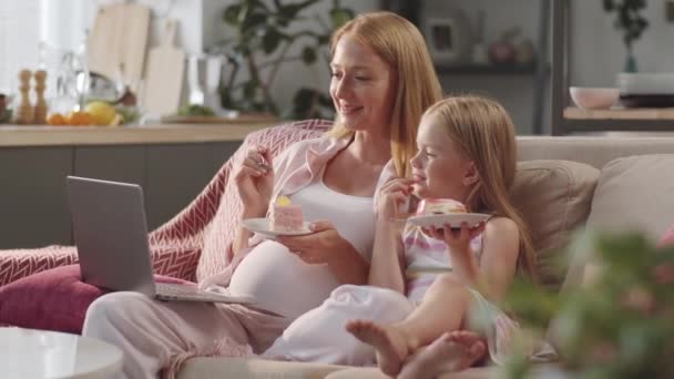 Χαρούμενο κοριτσάκι και η έγκυος μητέρα της κάθονται μαζί στον καναπέ στο σπίτι, τρώγοντας γλυκά κέικ, βλέποντας ταινία στο διαδίκτυο, κουβεντιάζοντας και γελώντας - Πλάνα, βίντεο