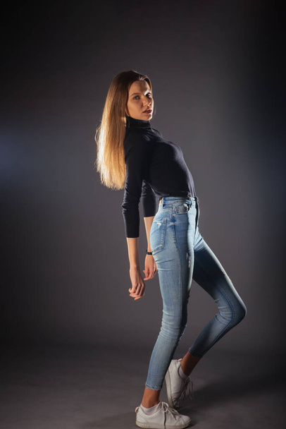 Side full-body portrait of a young female model in black top and jeans posing with one leg in front - Foto, afbeelding