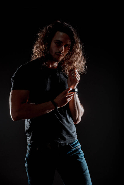 Cool young man dressed in black top, black jeans  posing in dark studio - Photo, Image