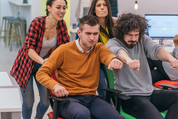 Team building and office fun.Four young cheerful businesspeople in smart casual wear having fun while racing on office chairs and smiling. - Photo, Image