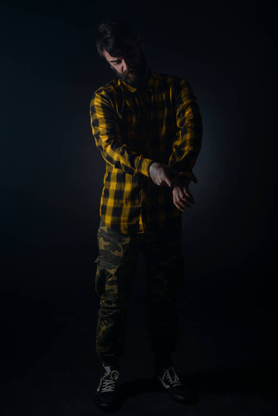 Dark image of a bearded man with tattoos fixing his buttons on his yellow and black checkered shirt - Fotoğraf, Görsel