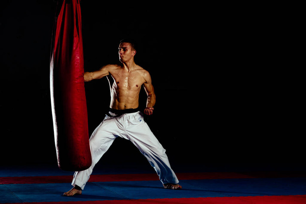 Luchador muscular en un guardia de combate junto al saco de boxeo aislado sobre fondo negro
 - Foto, Imagen