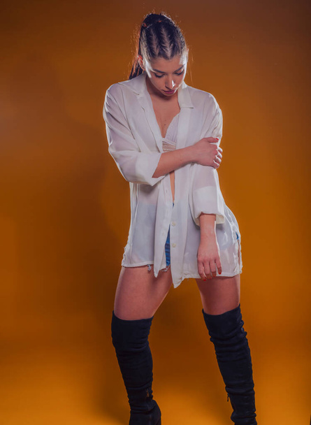Young woman wearing  stylish outfit with a white linen see-through shirt and black boots in a studio over an orange background - Foto, Imagem