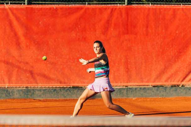 Nuori kunnon nainen pelaa tennistä ulkona oranssilla tenniskentällä aikaisin aamulla. - Valokuva, kuva