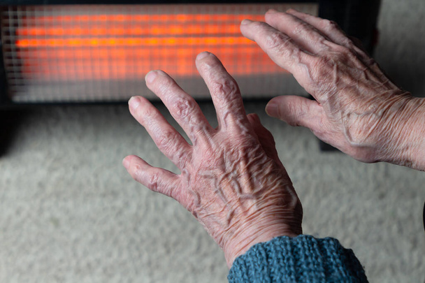 Vanhempi nainen lämmittää kätensä infrapunalämmittimen päälle. Kylmän sään turvallisuus vanhemmille aikuisille - Valokuva, kuva