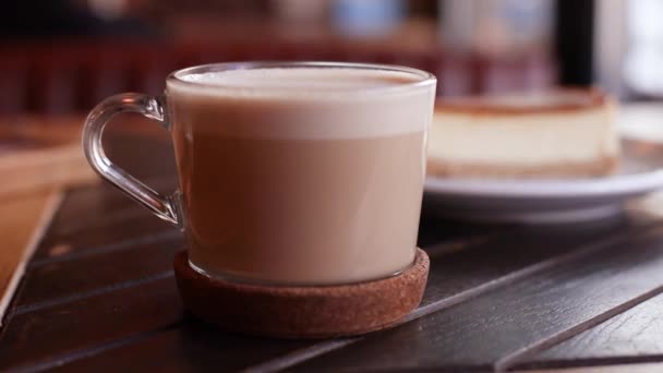 Primer plano de la taza de vidrio de café con leche caliente con un pastel de queso en el plato en la mesa - Metraje, vídeo