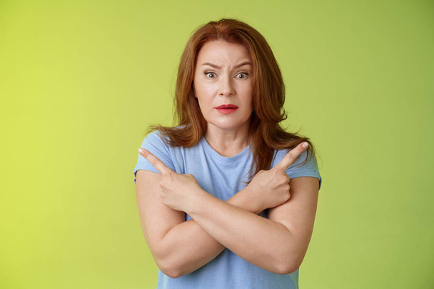 Help me choose. Worried unsure doubtful cute redhead middle-aged silly woman asking advice cross arms body pointing sideways left right look concerned camera make difficult decision green background - Photo, Image