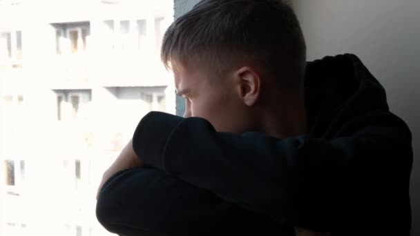 Θλιβερό έφηβο αγόρι με μαύρη κουκούλα κάθεται στο περβάζι του παραθύρου και κοιτάει έξω από το παράθυρο. Κοινωνική απόσταση κατά τη διάρκεια της καραντίνας Covid 19 Coronavirus στο σπίτι. Βίντεο 4k - Πλάνα, βίντεο