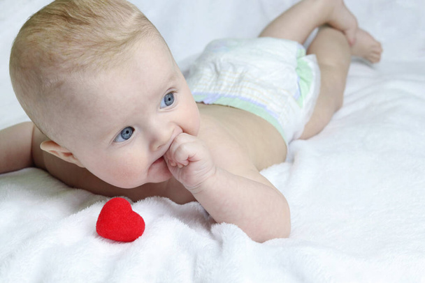 Close-up van een rood hart en een schattige baby liggend op een witte handdoek. Kauw en zuig aan je vingers. Kopieer ruimte - kiespijn concept, eerste tanden groeien, kwijlen - Foto, afbeelding