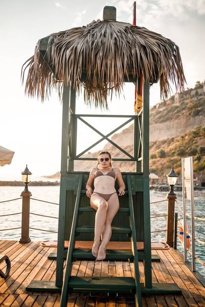 Vonzó fiatal nő bézs bikini fürdőruhában napnyugtakor ül egy faházban a tenger mellett. - Fotó, kép