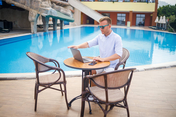 Sikeres, jóképű, napszemüveges üzletember egy medencénél ülő laptopnál dolgozik. Távoli munka. Szabadúszó - Fotó, kép