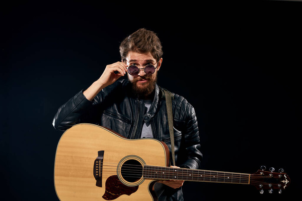 Ένας άντρας με μια κιθάρα στα χέρια του δερμάτινο μπουφάν μουσική παράσταση ροκ σταρ μοντέρνο στυλ σκούρο φόντο - Φωτογραφία, εικόνα