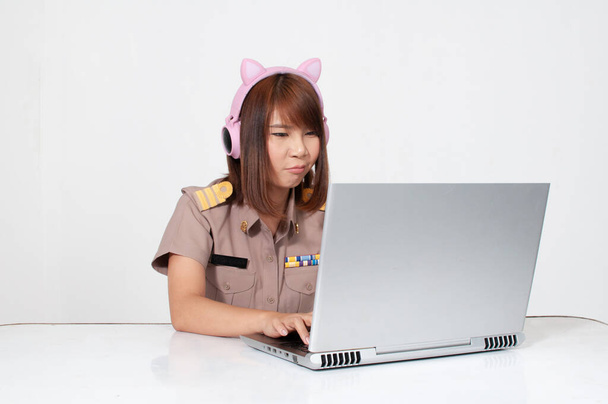 Insegnante thai donna in uniforme seduta con sfondo bianco utilizzando computer con cuffie auricolari rosa gatto - Foto, immagini