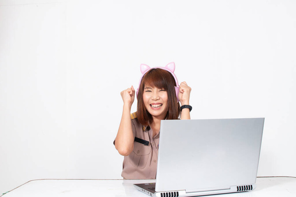 Female thai teacher in uniform sitting with white background using computer with pink cat ears headphone doing cheer up pos - Photo, Image
