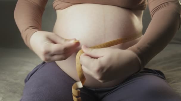 mujer embarazada joven sentada en el sofá, midiendo el vientre con cinta métrica en el interior - Imágenes, Vídeo