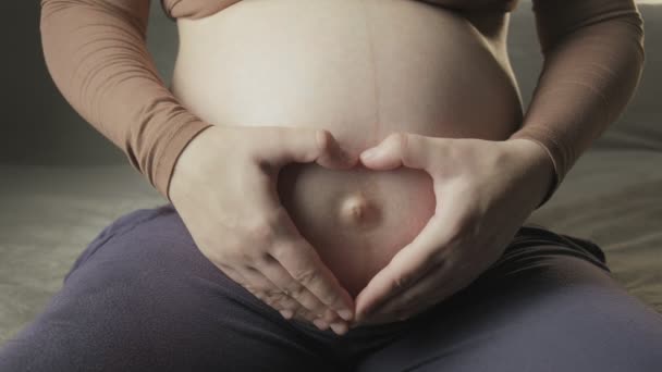 νεαρή έγκυος γυναίκα χαϊδεύοντας μεγάλη κοιλιά στον καναπέ, κάνοντας σχήμα καρδιάς με τα χέρια - Πλάνα, βίντεο