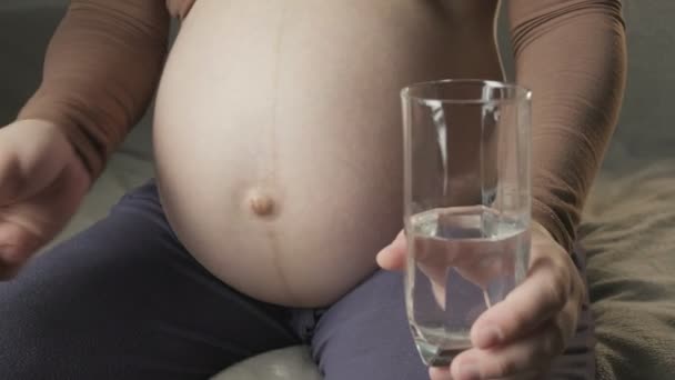 young pregnant woman sitting on couch, taking medicine, drinking glass of water - Materiał filmowy, wideo