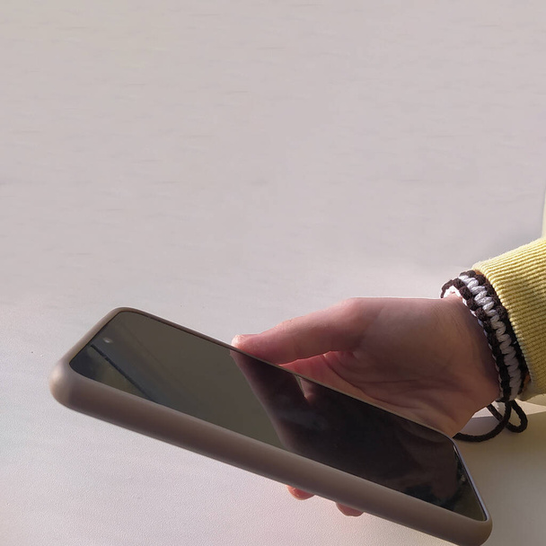 El adolescente tiene un teléfono móvil en la mano. La mano de una chica con un teléfono sobre un fondo claro. Pulsera hecha a mano, decoración. - Foto, imagen