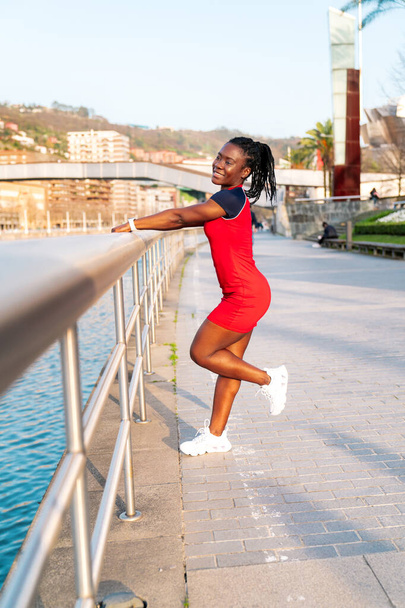 model girl with black skin and afro style hair posing smiling holding on to the railing of a bridge by the river of a city in a beautiful sunset with warm colours in a red dress - Фото, изображение