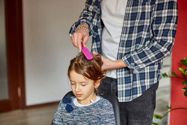 Vader kamt, poetst het haar van zijn dochter thuis, vader en dochter glimlachen, familiemomenten, brengt tijd samen door. - Foto, afbeelding