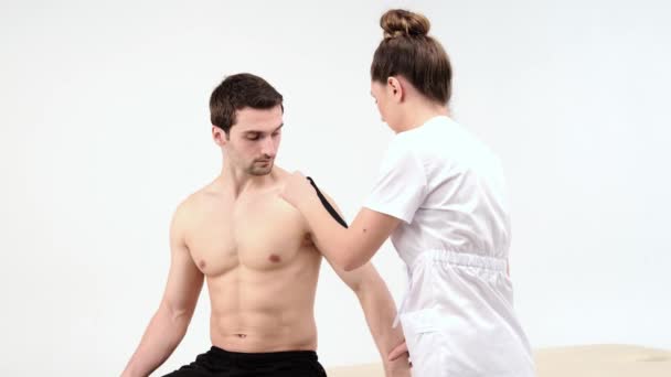 Tratamiento de hombro con cinta de cinesio. Fisioterapeuta aplicando cinta elástica terapéutica a la lesión del hombro del paciente. 4K. Fondo claro - Imágenes, Vídeo