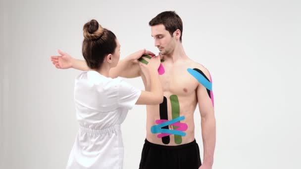 Schulterbehandlung mit Kinesiotape. Physiotherapeut legt elastisches Therapieband auf die Schulterverletzung des Patienten. 4K. Heller Hintergrund - Filmmaterial, Video