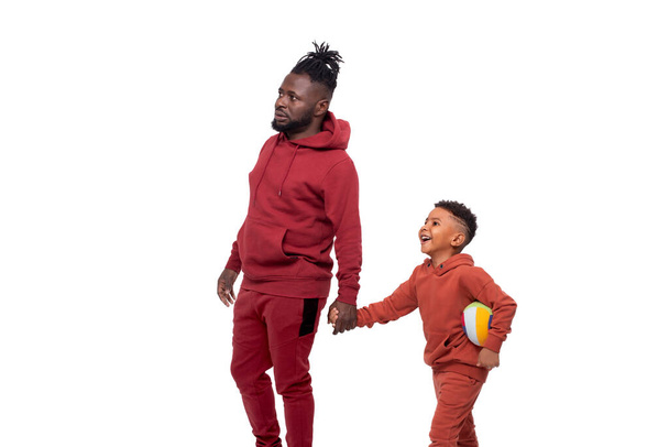 Pai e filho com bola, Africano de pele escura, vestindo os mesmos fatos de treino estão andando juntos em algum lugar. Conceito de valores familiares - Foto, Imagem