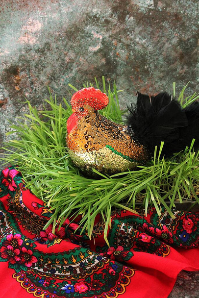 κόκορας διακοσμητικός, κόκορας με πούλιες σε φρέσκο πράσινο γρασίδι και με παραδοσιακό κόκκινο πανί, μαύρα φτερά στην ουρά του κόκορα - Φωτογραφία, εικόνα