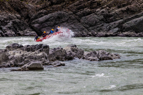 Une équipe de personnes rafting en gilets de sauvetage orange sur un bateau en caoutchouc de couleurs bleu et jaune le long d'une rivière de montagne sur le fond d'un rivage rocheux avec une forêt. - Photo, image