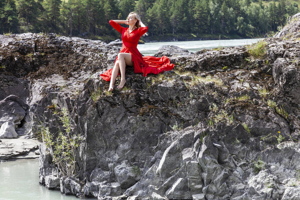 Blonde woman in red dress straightens hair with his hands, sits stretching her long legs on the rock stones on banks of Katun river against background of mountains on summer. Freedom and vacation. - Photo, Image
