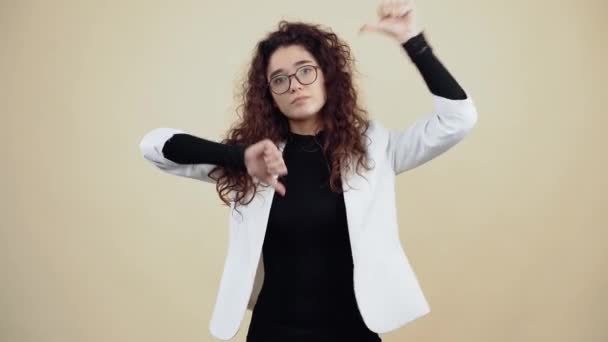 Bored young woman with curly hair shows her thumb down in denial, taster, ugly. Young hipster in gray jacket and white shirt, with glasses posing isolated on beige background in the studio. - Footage, Video