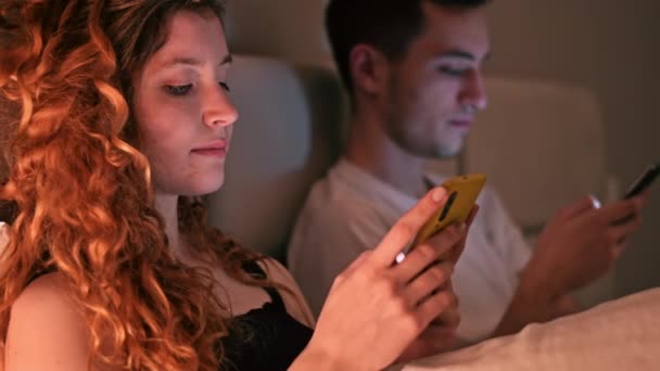 Νεαρό ζευγάρι που χρησιμοποιεί έξυπνα τηλέφωνα τη νύχτα στο κρεβάτι. Πολύχρωμο φως - Πλάνα, βίντεο